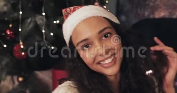 圣诞老人帽子里迷人的年轻微笑女子在智能手机上自拍坐在圣诞树附近的家中视频的预览图