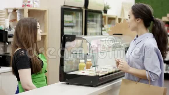 美丽的年轻女子带着纸袋在咖啡店里选择甜点友好的咖啡师穿着绿色围裙送着甜点视频的预览图