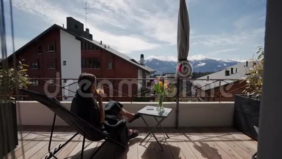 快乐的女人坐在家里的阳台上放松春天度假时在露台上喝橙汁视频的预览图