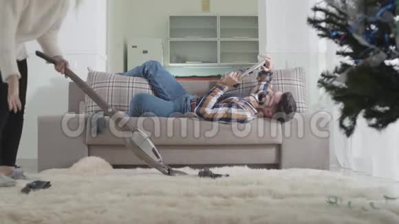 年轻的白种人金发女人在家里打扫卫生找到丈夫袜子和责骂男人懒汉长胡子躺在沙发上视频的预览图