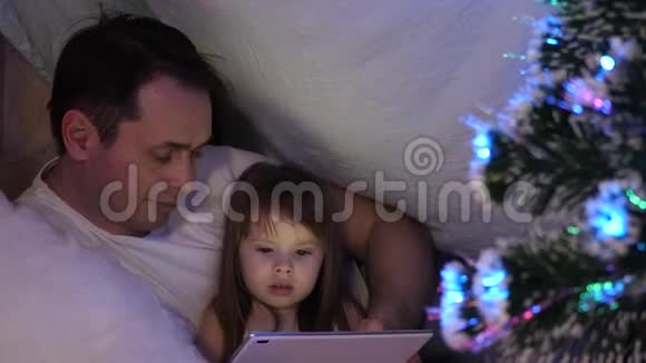 圣诞节晚上爸爸和女儿在平板电脑上玩和看动画片在一个带花环帐篷里的孩子房间里视频的预览图