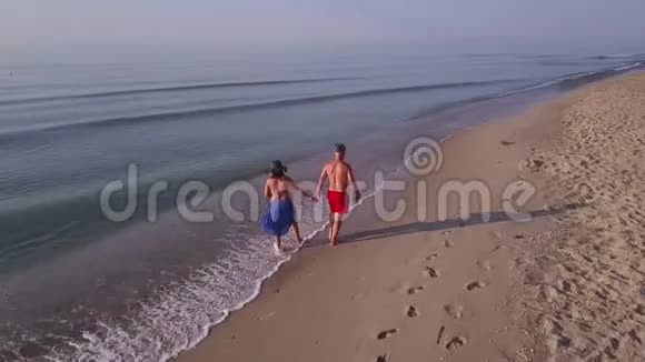 男人和女人在日落时沿着海滩散步追踪一对夫妇在海滩上玩得开心的空中镜头视频的预览图