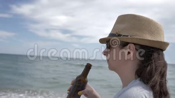 一个戴墨镜的女人从海边的瓶子里喝啤酒的肖像视频的预览图