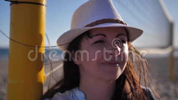 戴着帽子的幸福女人的肖像站在沙滩上的排球网附近看着镜头视频的预览图