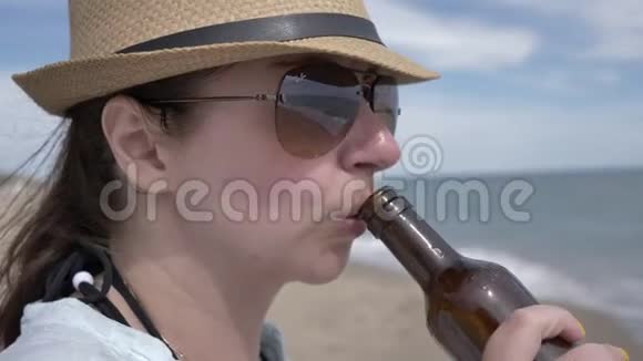 一个戴墨镜的女人从海边的瓶子里喝啤酒的肖像视频的预览图