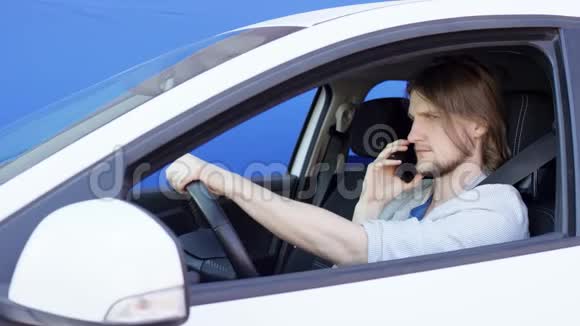 年轻人正在打电话而他正在驾驶一辆蓝色背景的白色汽车穿休闲夹克的男人视频的预览图