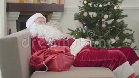 穿着圣诞老人服装睡在圣诞树背景的沙发上的老白种人精疲力竭的家伙休息视频的预览图