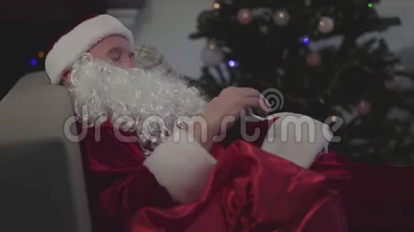 侧面肖像成熟的白人男子穿着圣诞老人服装睡在圣诞树的背景累了视频的预览图
