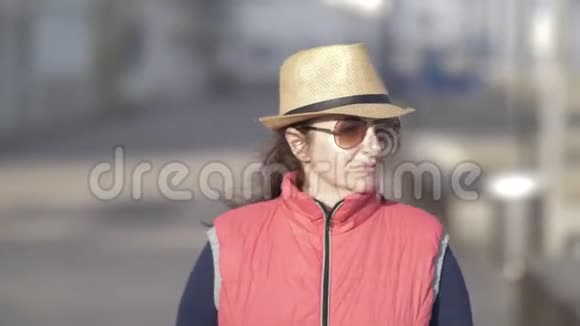 一位戴着帽子戴着墨镜的快乐旅行女人在风和日丽的天气下走在海边的大街上视频的预览图