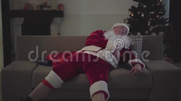 穿着圣诞老人服装戴着假白胡子的醉汉躺在家里的沙发上双腿分开视频的预览图