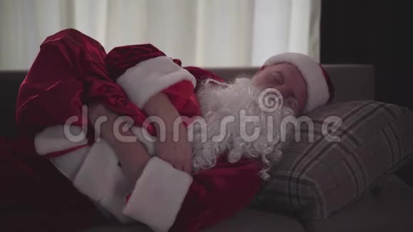 穿着圣诞老人服装睡在沙发上拿着带礼物的包疲惫不堪的高年级考卡西亚人精疲力竭的老人视频的预览图