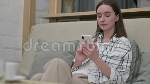 美丽的年轻女子坐在沙发上使用智能手机视频的预览图