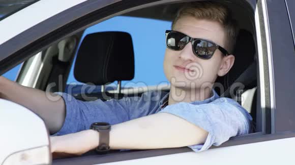一个黑发男人坐在蓝色背景的白色汽车里对着摄像头微笑他靠在车窗上他是视频的预览图
