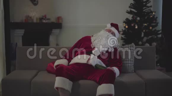 穿着圣诞老人服装睡在沙发上拿着瓶啤酒的白种人的肖像老头儿视频的预览图