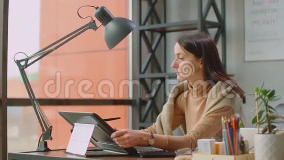 年轻的女人坐在她的办公桌前她在画画写作用钢笔与数字平板电脑手拿笔视频的预览图