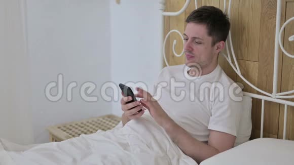 有魅力的年轻人坐在床上用智能手机发短信视频的预览图
