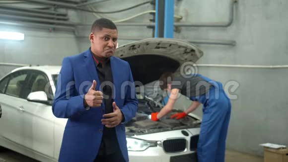 慢动作男人微笑并表现出良好的迹象机械师在后台修理一辆汽车视频的预览图