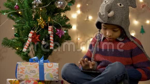 戴着大象帽的男孩在圣诞树附近玩智能手机视频的预览图