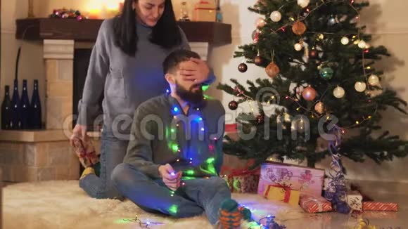 可爱的快乐年轻夫妇坐在房间地板上的圣诞树旁给男人送礼物的女人新的视频的预览图