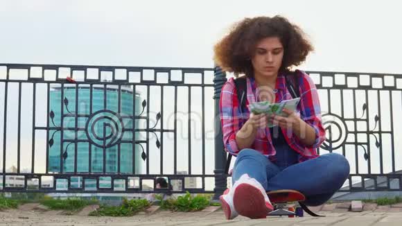 一个留着非洲发型的少女坐在城市街道上的滑板上数着欧元视频的预览图