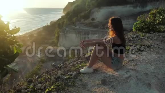年轻女子坐在岩石的背景上享受着大自然的美丽女人看着海洋日落背景视频的预览图