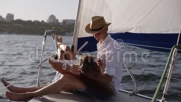 年轻浪漫的一对夫妇坐在游艇上拥抱船头长发黑发女孩躺在男朋友膝盖上视频的预览图