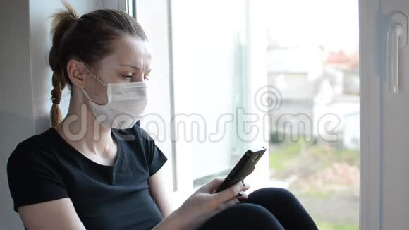 一个戴着医用口罩的年轻女孩坐在靠近窗户的窗台上看着智能手机视频的预览图