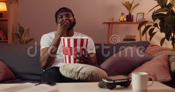 在家里坐在沙发上看无聊电影的小胡子家伙戴帽子的英俊非洲男人吃爆米花视频的预览图