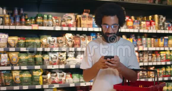 戴眼镜的长胡子的帅哥在用智能手机购物时带着篮子30岁的成熟男人在检查名单视频的预览图
