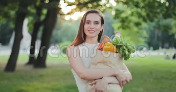 欢快的女孩一边拿着带食物的纸袋一边看镜头的肖像美丽的年轻女子微笑着摆姿势视频的预览图