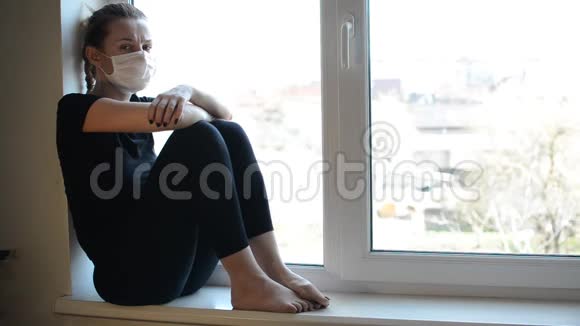 戴着医用口罩的年轻女孩坐在靠近窗户的窗台上视频的预览图