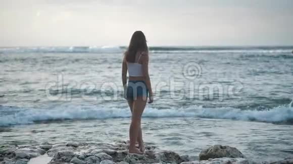穿着牛仔短裤的女孩在石滩上散步海浪拍打着海岸4k视频的预览图