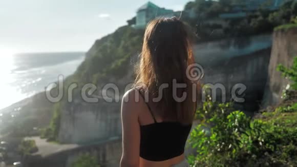 站在悬崖上看海洋的女孩的肖像她的头发是吹风的日落背景4k视频的预览图