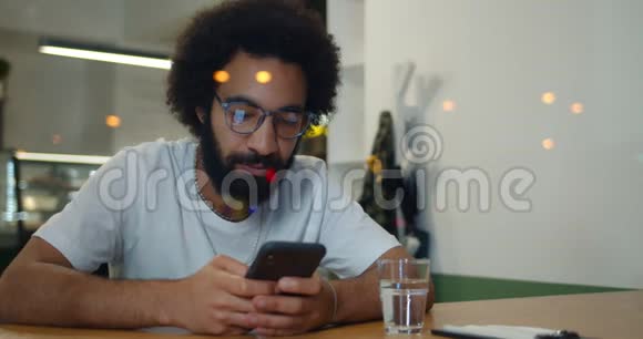 一个30多岁的大胡子他用智能手机坐在咖啡馆里微笑戴眼镜和T恤衫的快乐男人在聊天视频的预览图