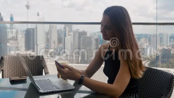 漂亮的女人坐在一家有笔记本电脑的咖啡馆里点击摩天大楼背景4k的智能手机视频的预览图