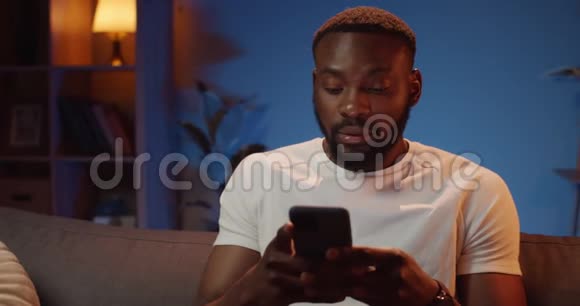 非洲年轻人使用智能手机和做出惊讶的面部表情的正面视图坐在沙发上的帅哥视频的预览图