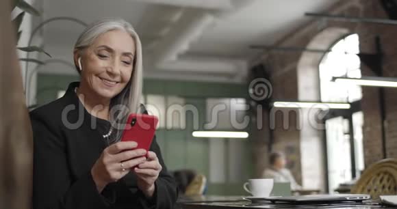 成熟的微笑女商人使用她的现代智能手机和看屏幕的正面视图快乐的现代老人视频的预览图