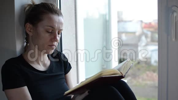 一个年轻的女孩坐在靠近窗户的窗台上看书视频的预览图