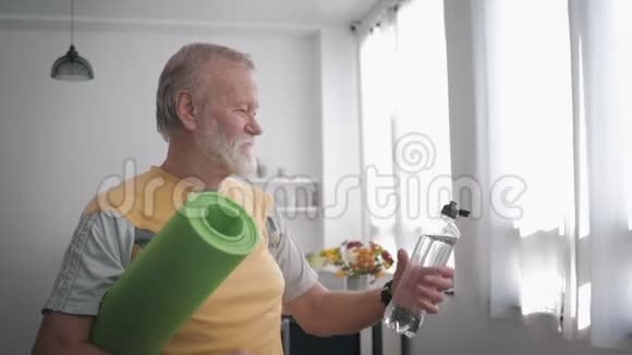 运动爷爷喝干净的水来保持健康手里拿着瑜伽垫站在室内视频的预览图