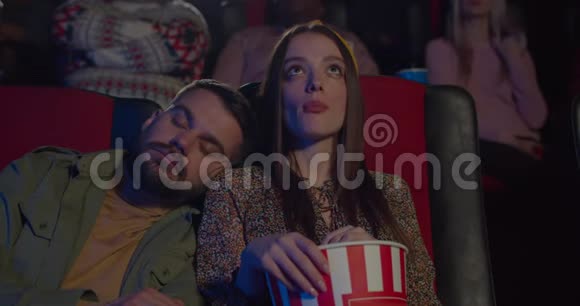 在无聊的电影中英俊的胡子男人睡在她女朋友的肩膀上年轻漂亮的女孩吃爆米花视频的预览图