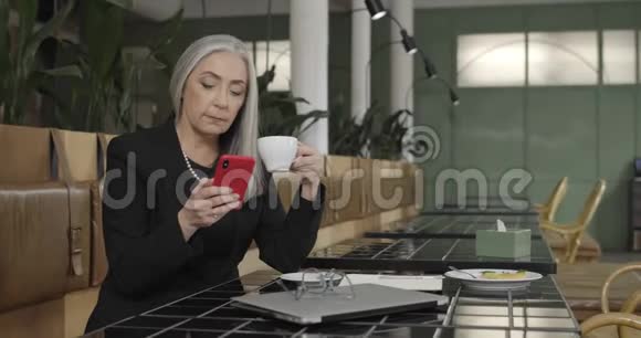 休息时成熟优雅的女商人坐在咖啡桌旁老年女性使用智能手机和视频的预览图