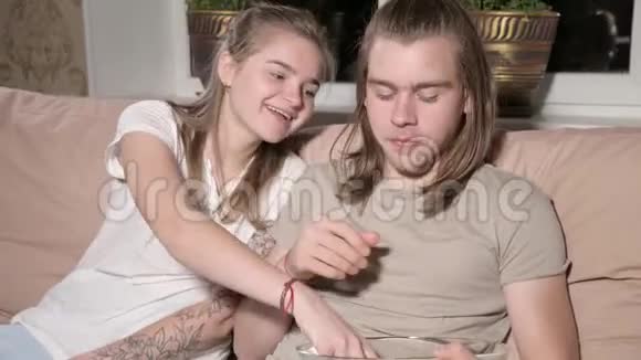 年轻快乐的夫妇正坐在沙发上女人把爆米花塞进男人嘴里视频的预览图