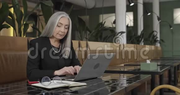 优雅的老女士使用笔记本电脑和打字同时看着屏幕和微笑美丽的中年妇女视频的预览图