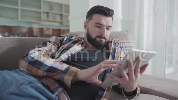 浓白种人留着胡须的男人躺在沙发上用平板电脑的特写肖像放松的家伙上网视频的预览图