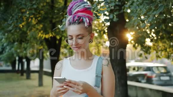 时尚女士在公园外面用智能手机微笑着享受社交媒体视频的预览图