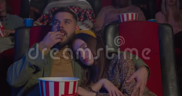 在电影院看电影时年轻女孩靠在丈夫肩膀上睡觉的正面景色吃爆米花和拥抱的大胡子视频的预览图