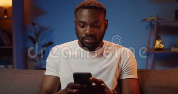 坐在沙发上的非洲男子留胡子身穿白色t恤打字留言帅哥在用电话视频的预览图