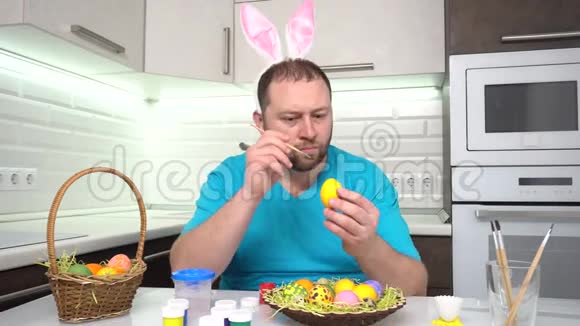 复活节快乐一个穿着兔子耳朵蓝色t恤的男人在厨房里画准备复活节的鸡蛋视频的预览图