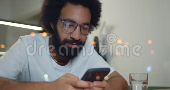 戴眼镜的帅哥在咖啡馆里坐在桌子旁用他的现代手机近距离观看胡须男打字视频的预览图
