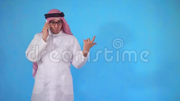 愤怒的阿拉伯男人冲动地站在蓝色背景上打电话视频的预览图